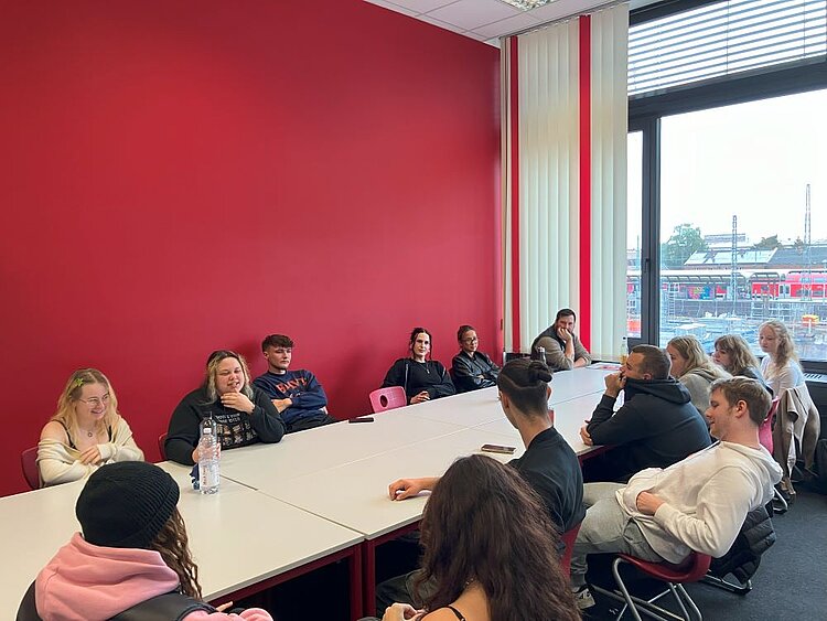 Wer sind die „Neuen“? – Kennenlernen der neuen Studierenden und Schüler*innen in Bamberg Eine Gruppe von jungen Erwachsenen sitzt in einem hellen Raum mit einer großen Fensterfront und einer roten Wand. Die Personen sind um einen langen, weißen Tisch herum platziert und wirken aufmerksam oder im Gespräch. Einige lächeln, andere hören zu. Im Hintergrund sind Gebäude und ein Zug zu sehen, was auf eine städtische Umgebung hindeutet.