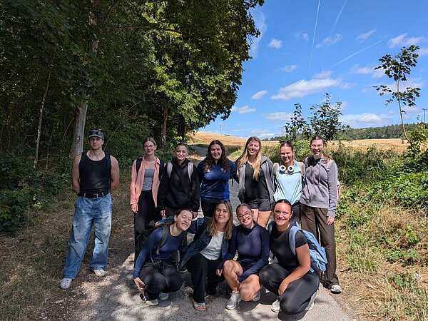 Eine Gruppe von elf Personen posiert auf einem Waldweg. Die meisten lächeln in die Kamera, während sie in einer lockeren Formation stehen. Im Hintergrund sind Bäume und eine offene Landschaft zu sehen.