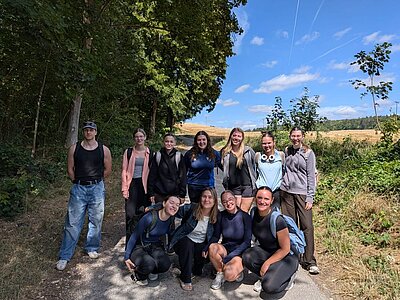 Eine Gruppe von elf Personen posiert auf einem Waldweg. Die meisten lächeln in die Kamera, während sie in einer lockeren Formation stehen. Im Hintergrund sind Bäume und eine offene Landschaft zu sehen.