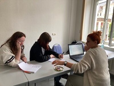 Zu sehen sind drei Schülerinnen, die mit Notebooks und Tablets an einem Tisch in einem Klassenraum sitzen und an einem Projekt arbeiten.