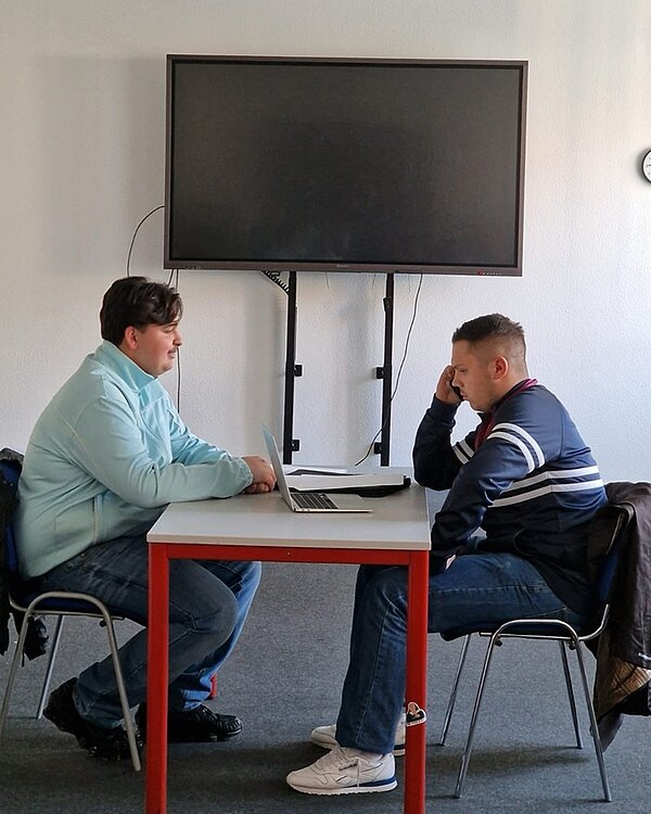 Theaterbesuch und Workshop Zwei männliche Auszubildende sitzen sich im Workshop in einem großen Raum an einem Tisch gegenüber.