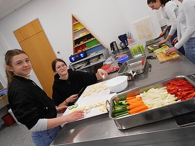 Mehrere Jugendliche bereiten in einer Schulküche Gemüse und Eier zu. Auf dem Tisch liegen farbenfrohe Gemüsestreifen, während zwei Mädchen vorne lächeln und mit einem Messer arbeiten.