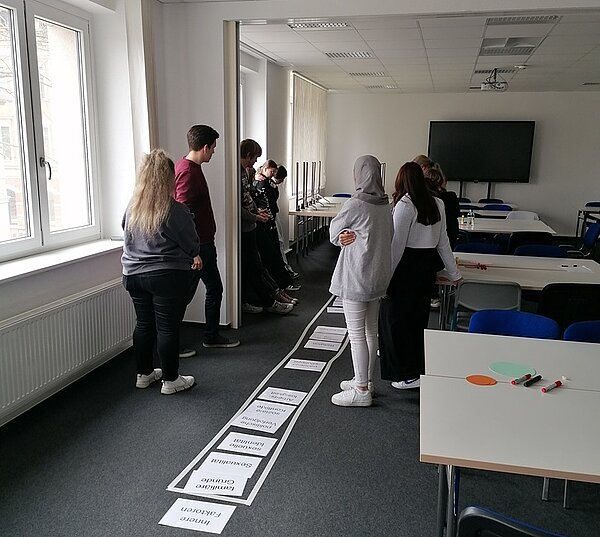 Die Auszubildenden stehen in einem Klassenraum an der Fensterfront links und rechts, auf dem Boden liegen Informationsblätter zum Workshop.