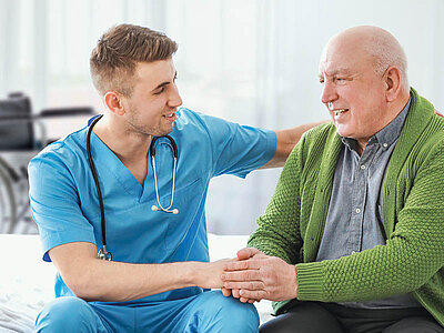 Eine medizinische Fachkraft in blauen OP-Kleidung und einem umgehängten Stethoskop tröstet einen älteren Patienten in einem grünen Cardigan während einer Konsultation.