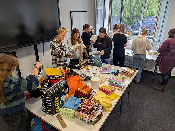 Mehrere Azubis stehen um einen Tisch, der mit buntem Papier, Stoffen, Taschen und Bastelmaterialien bedeckt ist. Einige schneiden oder falten Materialien, andere unterhalten sich. Im Hintergrund sind große Fenster mit Blick auf Bäume und eine Mauer. Die Szene wirkt wie ein gemeinsames Bastel- oder Kreativprojekt.