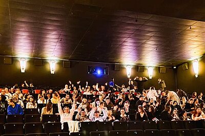 In einem fast vollbesetzten Kinosaal heben einige Besucher die Arme hoch, andere sind gerade dabei, sich zu setzen. Die Saalbeleuchtung ist noch an. Im oberen mittleren Bereich der hinteren Wand ist das blaue Leuchten des Filmprojektors zu sehen.