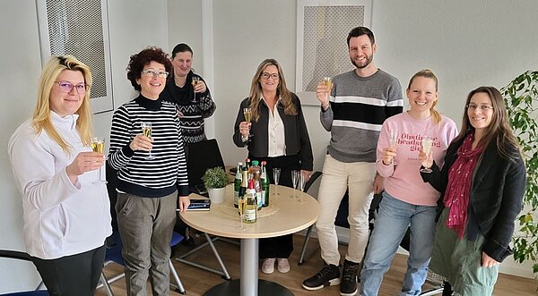Eine Gruppe von sieben Personen steht in einem Raum und hält Sektgläser in die Höhe. Sie lächeln und scheinen eine Feier oder ein besonderes Ereignis zu genießen. Auf dem Tisch in der Mitte stehen mehrere Sektflaschen und Gläser. Die Atmosphäre ist fröhlich und entspannt.