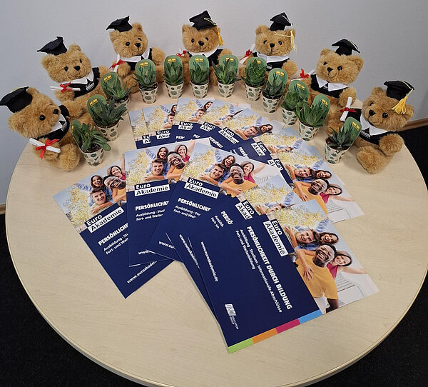 Mehrere braune Teddybären mit schwarzer Absolventenmütze und weißem Kragen tragen kleine Töpfe mit grünen Pflanzen. Sie sind auf einem runden Holztisch arrangiert, um den herum Flyer der Euro Akademie mit dem Motto ‚Persönlichkeit durch Bildung‘ ausgelegt sind.