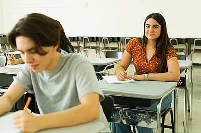 Zwei Personen sitzen an Einzeltischen in einem Klassenraum. Die Person im Vordergrund trägt ein helles T-Shirt und hält einen Stift in der rechten Hand. Die Person im Hintergrund trägt eine gemusterte, kurzärmelige Bluse und schreibt auf ein Blatt Papier. Im Raum stehen mehrere leere Stühle und Tische, der Boden ist hell und der Hintergrund unscharf.
