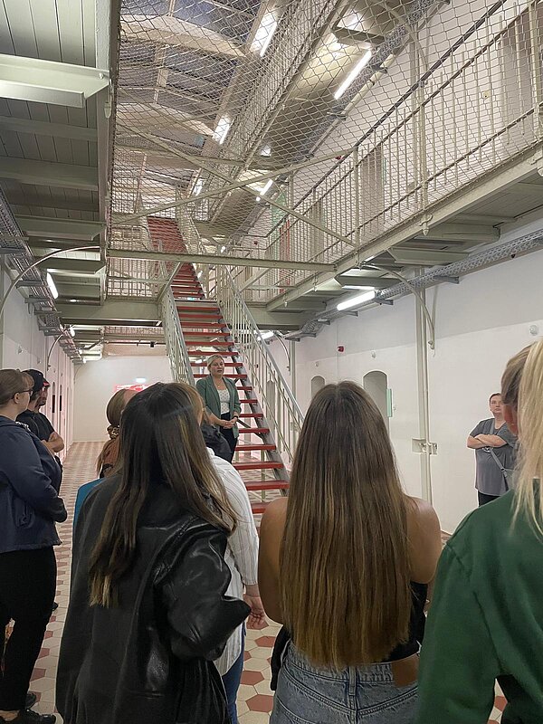 Gruppe von Besucherinnen und Besuchern bei einer Führung im Inneren des Kaßberg-Gefängnisses. Sie stehen vor einer roten Treppe, über ihnen verlaufen Gittergänge und Netze.