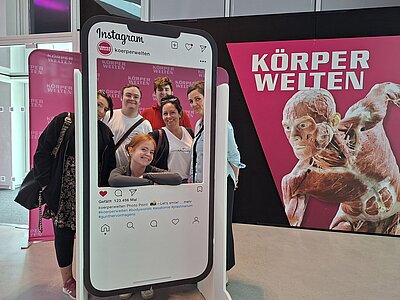 Eine kleinere Gruppe von Personen posiert fröhlich hinter einem großen Instagram-Fotorahmen in der Ausstellung "Körperwelten". Im Hintergrund ist ein weiteres Plakat mit einer anatomischen Figur sowie der Schriftzug "KÖRPERWELTEN" in Weiß auf pinkem Grund zu sehen.