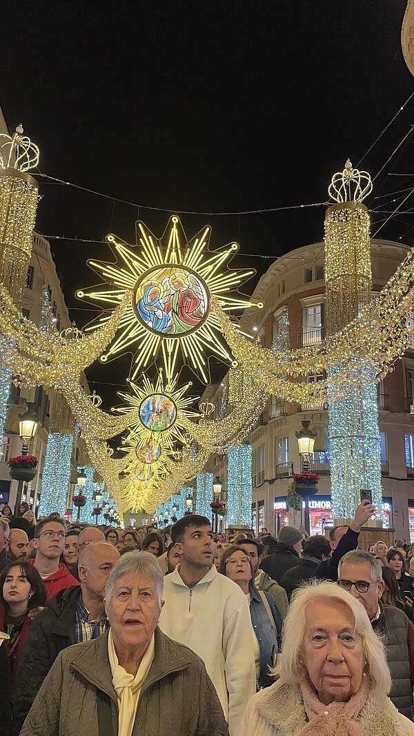 Menschenmenge in einer Einkaufsstraße bei Nacht, die nach oben zu einem spektakulären Weihnachtslichtbogen mit goldenen Sonnenmotiven, Engeln und funkelnden Lichterketten schaut.