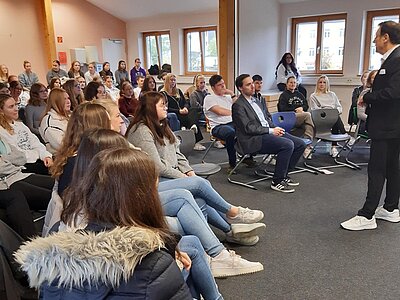 Ein Raum voller Menschen, die auf Stühlen sitzen und einem Vortrag oder einer Präsentation zuhören. Ein Mann im Anzug steht vorne und scheint die Präsentation zu halten. Der Raum hat große Fenster, die viel natürliches Licht hereinlassen.