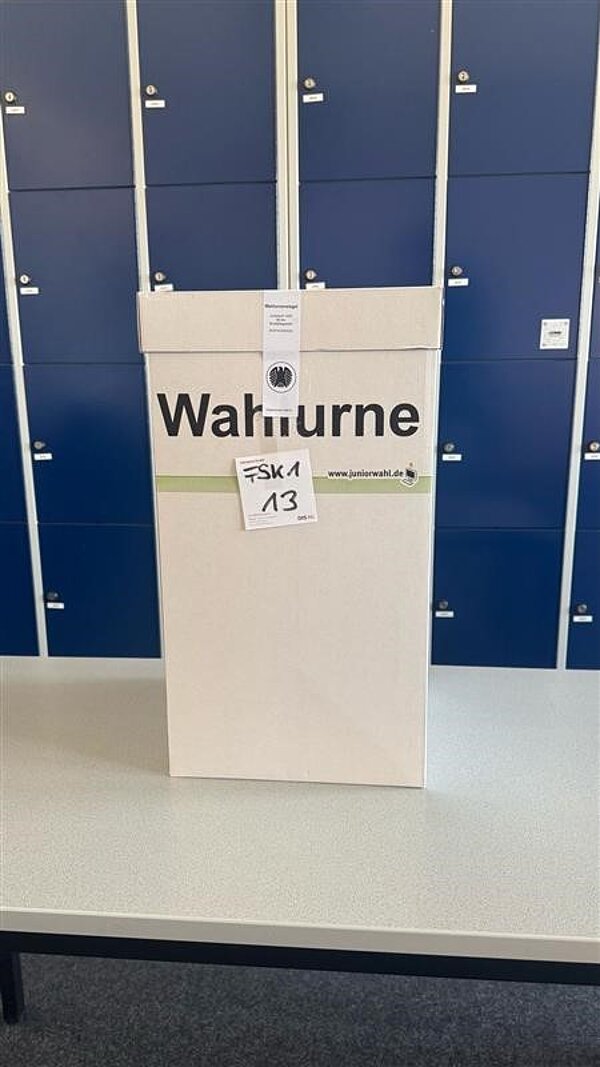 Testwahlen an der Euro Akademie Aschaffenburg am 19. Februar 2025 Eine weiße Wahlurne mit der Aufschrift „Wahlurne“ steht auf einem Tisch. Im Hintergrund sind blaue Schließfächer zu sehen.