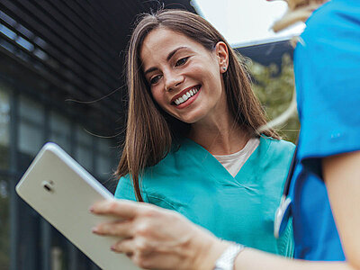 Eine lächelnde Krankenschwester in grüner Uniform hält ein Tablet in den Händen und blickt auf den Bildschirm. Sie steht neben einer anderen Person in blauer Kleidung, die teilweise sichtbar ist. Der Hintergrund zeigt ein modernes Gebäude mit Glasfassade.