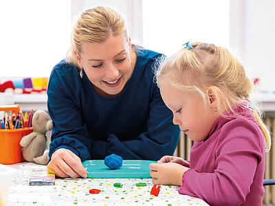 Eine Frau und ein kleines Mädchen sitzen an einem Tisch und basteln gemeinsam. Die Frau trägt ein dunkles Oberteil und lächelt freundlich, während sie dem Mädchen hilft. Das Mädchen, mit blonden Haaren und einem rosa Pullover, konzentriert sich auf das Bastelprojekt vor ihr. Auf dem Tisch liegen bunte Bastelmaterialien wie Knetmasse, Schere, Klebestift und verschiedene Papiere. Im Hintergrund sind ein Behälter mit Buntstiften und ein kleiner Stofftier-Hase zu sehen. Beide wirken glücklich und engagiert. Die Szene vermittelt eine warme und kreative Atmosphäre.