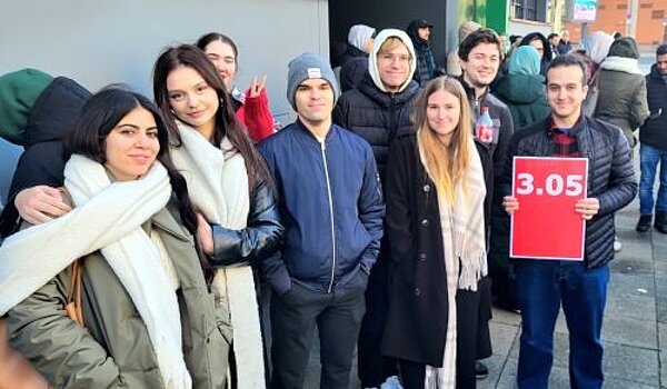 Euro Akademie und Euro-Schulen on Fire! Eine Gruppe von neun Personen posiert draußen in einer Menschenmenge. Zwei Frauen im Vordergrund tragen weiße Schals. Ein Mann in der Gruppe hält ein Schild mit der roten Aufschrift „3.05“. Alle Personen tragen Winterkleidung, darunter Jacken und Mützen. Weitere Menschen sind im Hintergrund zu sehen, ebenfalls warm angezogen.