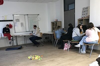 Ein Klassenzimmer oder Seminarraum mit mehreren Auszubildenden, die entweder zu zweit oder alleine an Tischen sitzen und konzentriert schreiben. Im Hintergrund hängt an einer Wand eine Magnettafel, an der mehrere beschriebene Zettel und ein großes Plakat hängen. Auf dem Boden in der Mitte des Raumes liegen viele bunte, beschriftete Zettel auf einem Haufen.