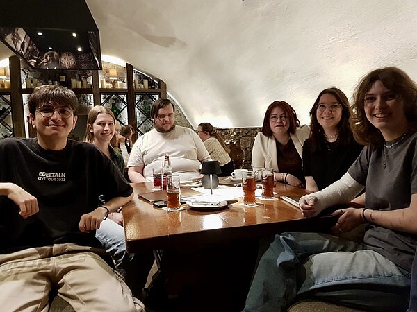 Alumnitreffen 2025 Sechs ehemalige Auszubildende der Euro Akademie Aschaffenburg sitzen an einem Tisch im Restaurant und posieren lächelnd für ein Gruppenfoto. Vor ihnen liegen Speisekarten, Gläser und eine Tasse mit verschiedenen Getränken, Besteck und eine kleine Tischlampe.