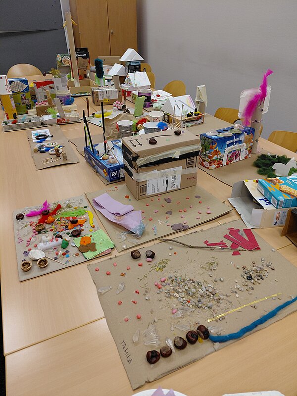 Verschiedene flache Collagen aus Karton liegen auf einem Tisch, verziert mit Steinen, Kastanien, Federn, bunten Perlen und Stoffstücken; dahinter stehen weitere dreidimensionale Kartonbauten und Bastelobjekte.