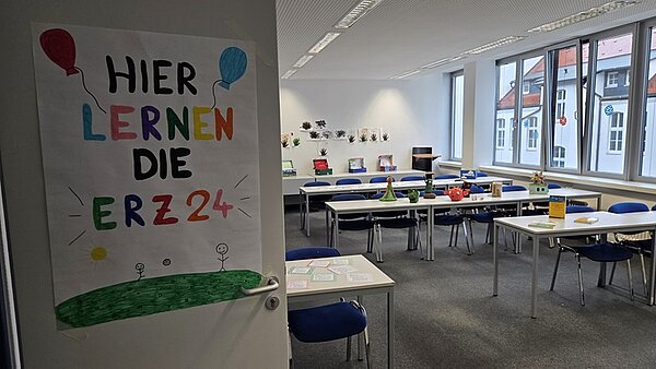Ein Klassenzimmer der Euro Akademie Erfurt mit weißer Decke, farbenfrohen Dekorationen, Tischen, Stühlen, einem Computermonitor, einem Drucker und einer Vase auf einem grauen Teppich. An der Tür das selbst gestaltete Bild "Hier lernen die ERZ 24".