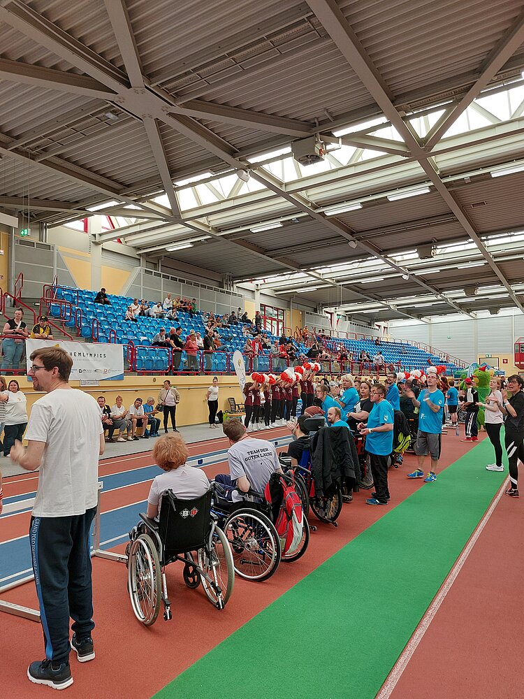 Teilnehmende und Zuschauer*innen versammeln sich bei einer inklusiven Sportveranstaltung in einer großen Leichtathletikhalle; mehrere Personen sitzen im Rollstuhl.