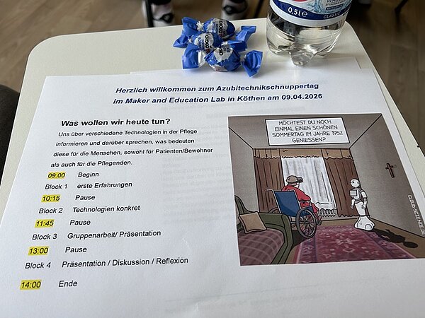 Im Vordergrund liegt ein Blatt Papier auf einem Tisch. Das Blatt trägt die Überschrift 'Herzlich willkommen zum Azubitechnikschnuppertag im Maker und Education Lab in Köthen am 09.04.2026'. Darunter folgt ein Tagesablauf mit Uhrzeiten und Programmpunkten, die in verschiedenen Farben hervorgehoben sind. Rechts auf dem Blatt befindet sich eine farbige Illustration: Ein humanoider Roboter steht neben einem Bett, in dem eine Person sitzt. Über dem Roboter steht eine Sprechblase mit dem Text: 'MÖCHTEST DU NOCH EINMAL DEINEN SCHÖNEN SOMMERFILM IM SAAL SEHEN?'. Auf dem Tisch liegen außerdem zwei Bonbons mit blau-weißer Verpackung und eine Wasserflasche.