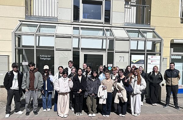 Die Gruppe der Auszubildenden posiert vor einem Gebäude mit Glastüren und -fenstern. Die Gruppe besteht aus Männern und Frauen, die meisten tragen legere Kleidung wie Jacken und Mäntel. Das Gebäude hat Schilder in deutscher Sprache und die Hausnummer 71. Das Wetter scheint sonnig zu sein.