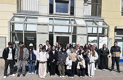 Die Gruppe der Auszubildenden posiert vor einem Gebäude mit Glastüren und -fenstern. Die Gruppe besteht aus Männern und Frauen, die meisten tragen legere Kleidung wie Jacken und Mäntel. Das Gebäude hat Schilder in deutscher Sprache und die Hausnummer 71. Das Wetter scheint sonnig zu sein.