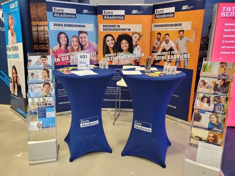 Rückblick auf die Jobmesse Leipzig Ein Messestand der Euro Akademie mit drei großen, bunten Bannern, die verschiedene Ausbildungs- und Studienangebote bewerben. Zwei mit blauen Tischhussen bedeckte Stehtische mit Flyern, Stiften und Infomaterialien stehen im Vordergrund. Rechts und links befinden sich Prospektständer mit weiteren Informationsbroschüren. Die Banner zeigen junge, lächelnde Menschen und Schlagworte wie „Zukunftsgestalter in die Berufswelt U21“, „Berufe für Herzensmenschen“ und „Sprungbrett für deine Karriere“.