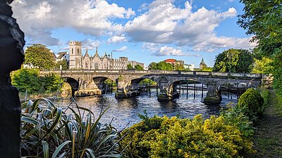 Eingerahmt in das Grün von Bäumen und Sträuchern sind die Salmon Weir Bridge und dahinter einige altertümliche Gebäude Galways zu sehen. Durch den überwiegend blauen Himmel ziehen einige dicke Kumuluswolken.