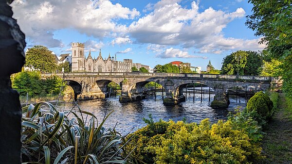 Willkommen zurück aus Galway! Eingerahmt in das Grün von Bäumen und Sträuchern sind die Salmon Weir Bridge und dahinter einige altertümliche Gebäude Galways zu sehen. Durch den überwiegend blauen Himmel ziehen einige dicke Kumuluswolken.