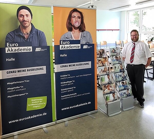 Freundlicher Mitarbeiter am Messestand der Euro Akademie Halle steht neben zwei großen Rollups und Regal gefüllt mit Broschüren und Flyern.