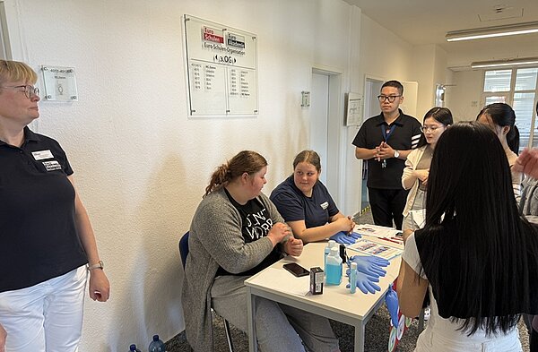 Tag der offenen Tür an der Euro Akademie Bitterfeld-Wolfen Besucherinnen des Tages der offenen Tür stehen um einen Tisch herum, an dem eine Auszubildende sitzt, stehend daneben eine Lehrkraft, beide in marineblauem Euro Akademie-T-Shirt, eine Besucherin sitzt auf dem Stuhl neben ihr, auf dem Tisch liegen Einmalhandschuhe in hellblau, Desinfektionsmittel etc.