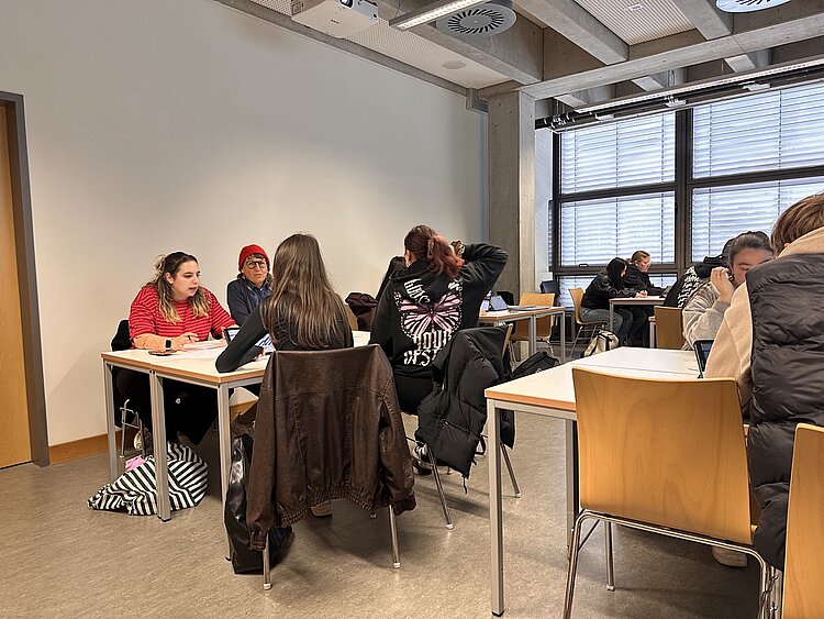 Studierende arbeiten in kleinen Gruppen an Tischen in einem Seminarraum und besprechen gemeinsam Aufgaben.