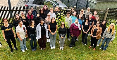 Große Gruppe von Jugendlichen und Erwachsenen versammelt sich für ein gemeinsames Foto auf einer Grünfläche rund um einen jungen Baum.