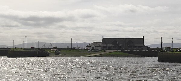 Ein Gebäude am Wasser mit einem kleinen Hafen oder Anleger, umgeben von einer grünen Landschaft.