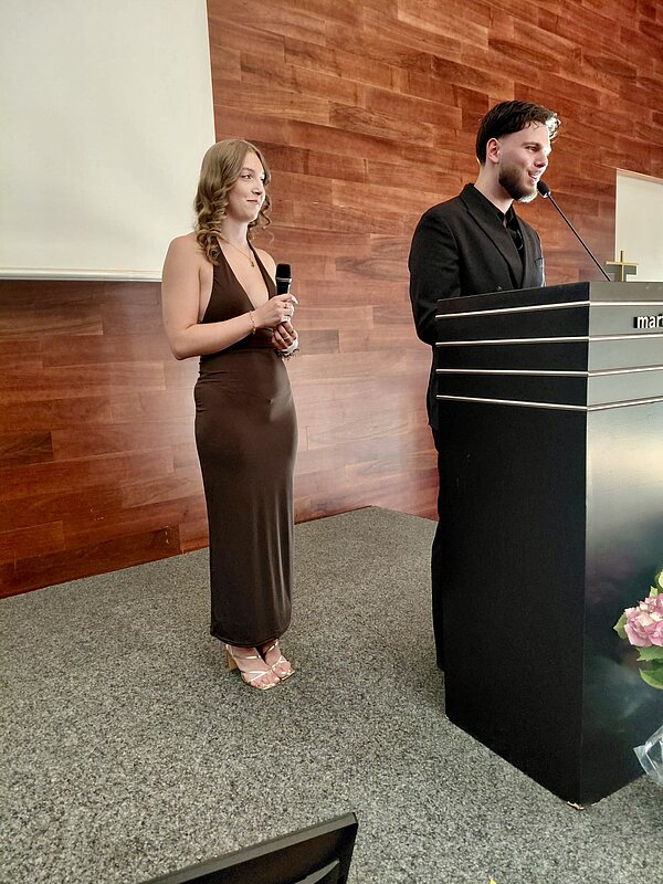 An einem Rednerpult steht ein junger Mann in schwarzem Hemd. Hinter ihm steht eine junge Frau in braunem Kleid mit einem Mikrofon in der Hand.
