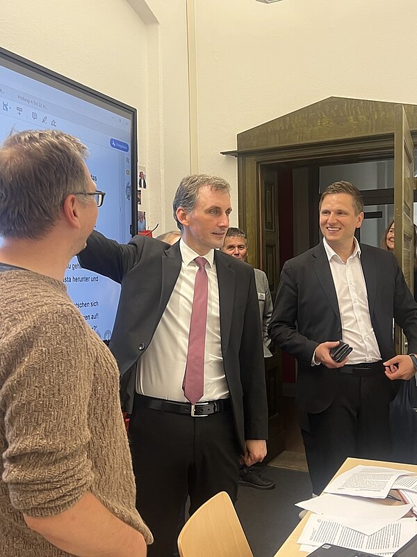 Ein Mann im Anzug mit rosa Krawatte steht in einem Raum und gestikuliert mit erhobenem Arm in Richtung eines großen Bildschirms, auf dem Text angezeigt wird. Ein weiterer Mann im Anzug hält ein Smartphone in der Hand. Im Vordergrund ist eine Person mit Brille und braunem Pullover von hinten zu sehen. Auf dem Tisch liegen mehrere Blätter Papier. Im Hintergrund sind weitere Personen teilweise sichtbar.
