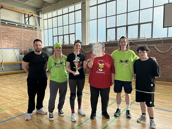 Siegerteam des Volleyballturniers in der Sporthalle: Sechs Personen posieren mit Pokal und Urkunde vor der Kletterwand.