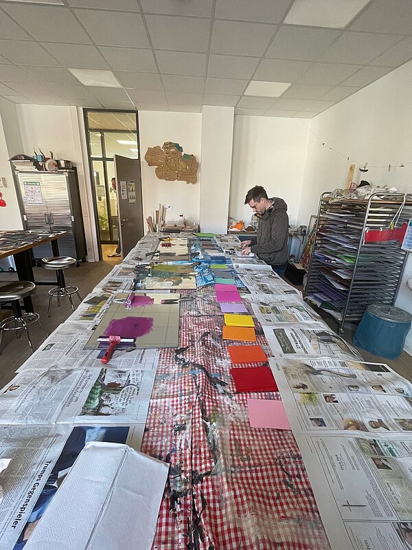 Ein heller Werkraum mit langem Arbeitstisch, der mit Zeitungen ausgelegt ist, darauf liegen Farbwalzen, Druckplatten und bunte Papierkarten.