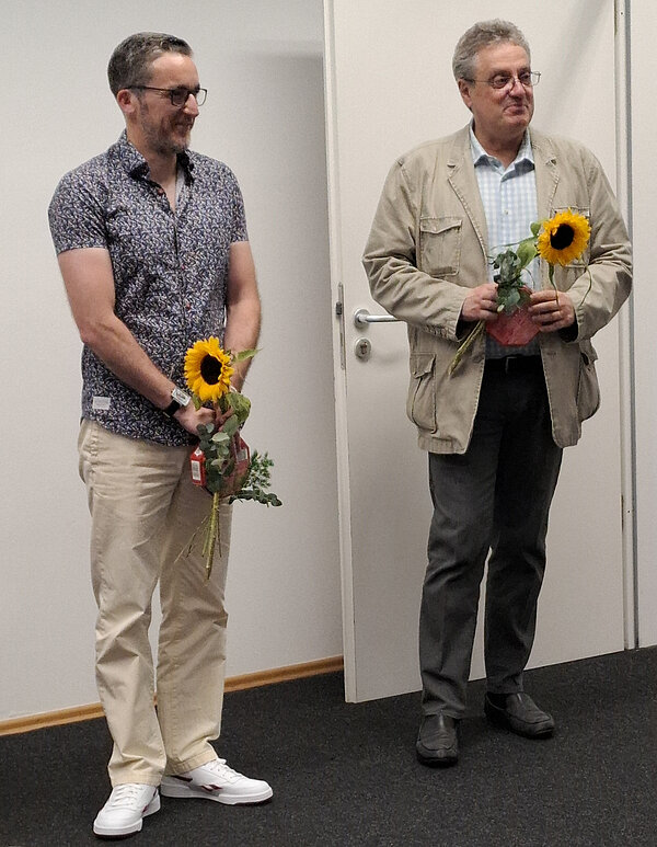 Zwei Männer stehen nebeneinander in einem Raum mit weißer Tür. Beide halten eine Sonnenblume und eine Pralinenschachtel in den Händen. Der Mann links trägt ein blaues kurzärmliges Hemd und helle Hosen, der Mann rechts ein beiges Sakko. Beide Männer tragen eine Brille.
