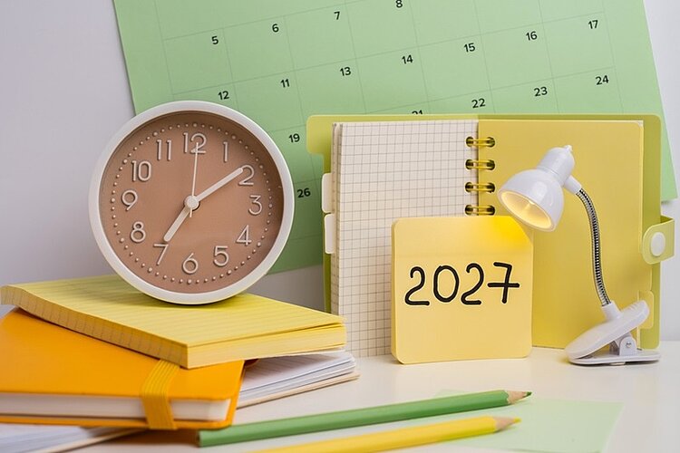 Auf einem Schreibtisch liegen mehrere gelbe und weiße Notizbücher, ein Bleistift, eine kleine Schreibtischlampe und eine analoge Uhr mit beigem Zifferblatt. Im Hintergrund ist ein Wandkalender zu sehen. Im Vordergrund steht ein aufgeschlagenes Notizbuch mit der handschriftlichen Jahreszahl '2027'.