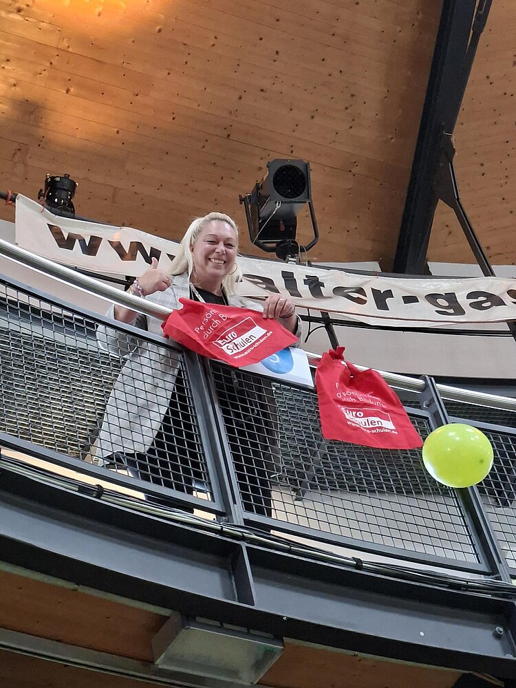 Eine lächelnde Frau steht auf einer umlaufenden Galerie in einem Veranstaltungsgebäude mit Holzkuppeldecke. Am Geländer hängen bunte Luftballons und rote Taschen. Die Aufnahme wurde von unten nach oben fotografiert.