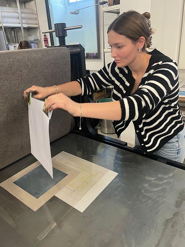 Eine Frau mit zusammengebundenen Haaren und schwarz-weiß gestreiftem Cardigan legt ein großes weißes Blatt Papier oder Stoff auf eine Fläche mit einer technischen Zeichnung auf – ein Arbeitsschritt an der Druckpresse in der Werkstatt.