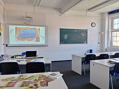 Ein Klassenzimmer mit weißen Tischen und blauen Stühlen; links vorne eine Leinwand mit dem ESO-Logo, rechts daneben eine Tafel mit den Worten "Herzlich Willkommen 2024."