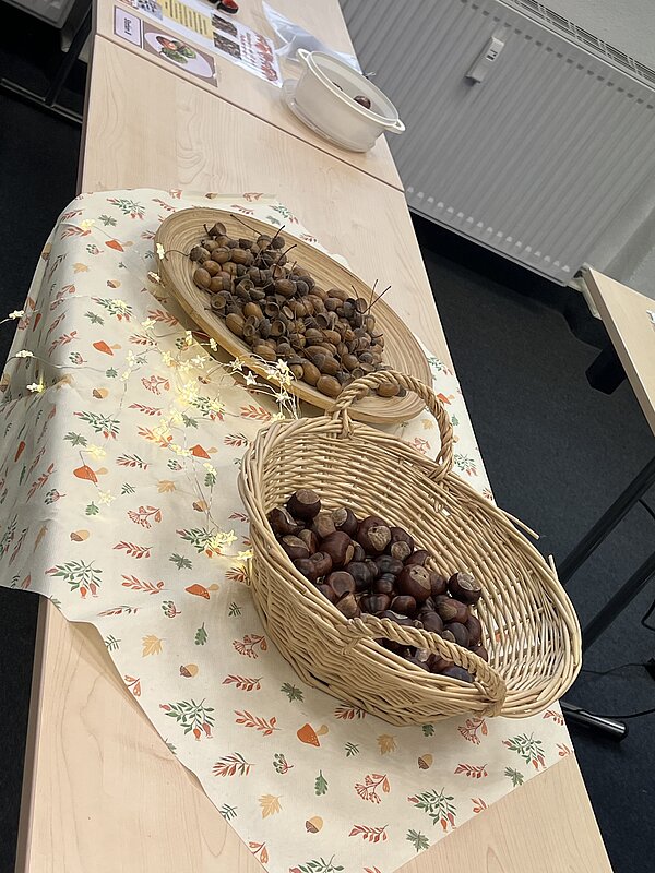 Auf einem herbstlich dekorierten Tisch stehen zwei Weidenkörbe, einer mit Kastanien und einer mit Eicheln auf Spießen, umgeben von Lichterkette und Infomaterial.
