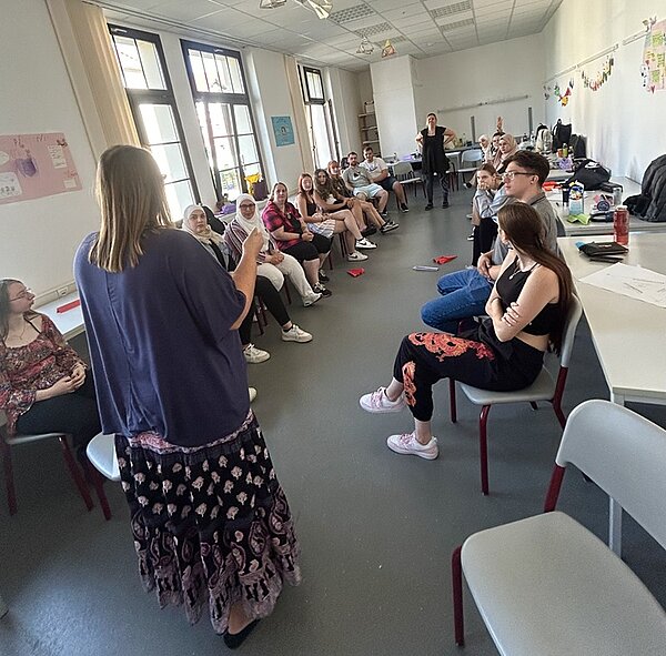 Schüler sitzen sich in einem Raum in zwei langen Reihen gegenüber. 2 Schülerinnen tragen ein Kopftuch, die Lehrerin spricht zur Klasse. 