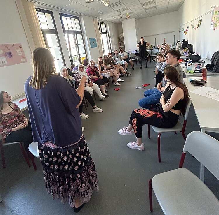 Schüler sitzen sich in einem Raum in zwei langen Reihen gegenüber. 2 Schülerinnen tragen ein Kopftuch, die Lehrerin spricht zur Klasse. 