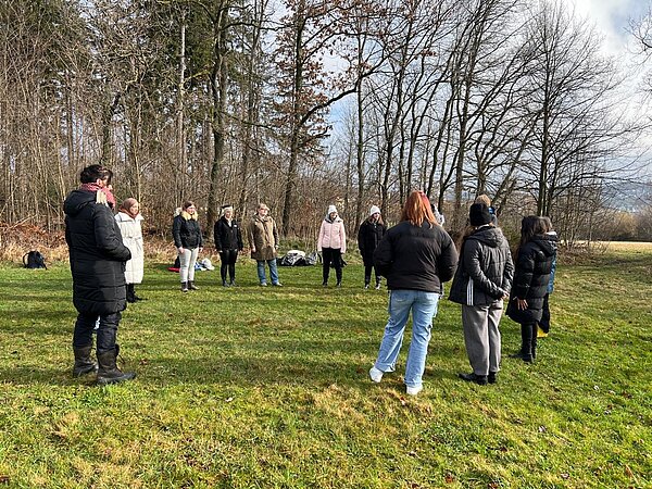 Die Schüler*innen stehen auf einer Wiese am Rand eines kleinen Waldstücks in einem Kreis und scheinen jemandem zuzuhören, der aber auf dem Bild von einer anderen Person verdeckt ist.